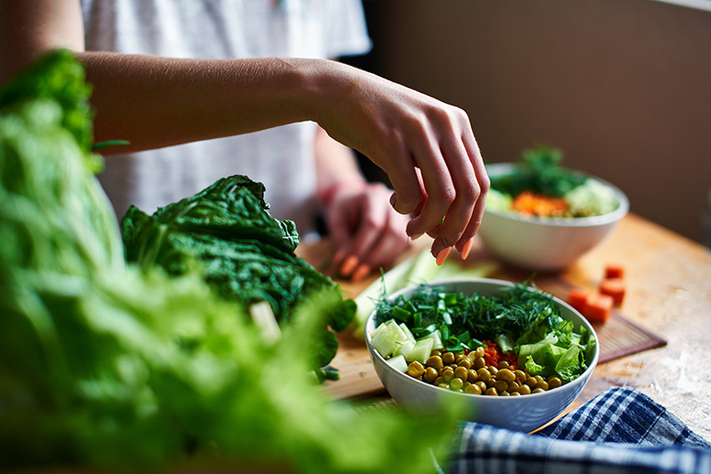 Female hand pours green onions in a bowl with green peas, cucumbers, carrots, lettuce and dill standing on a table
